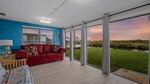 A spacious room with a red sofa, a wooden side table, and a view of a grassy area and water body through large glass doors. - Waterfront! 2BR w/Firepit & Grill! (Hudson)