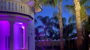 An outdoor patio area with a curved white building, palm trees, and tables and chairs. - Corte Vena Palace (Bianco)