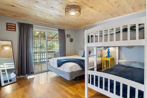 A room with a bunk bed, a small table with a lamp, a window with curtains, and a wooden ceiling.