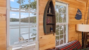 A cabin with large windows overlooking a lake, a wooden shelf with items, and a plaid-patterned bench. - Waterfront Home with Bunkhouse (Rocky Mount)