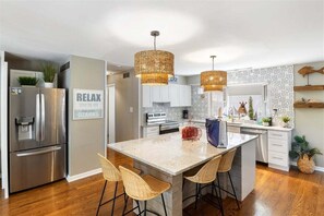 Une cuisine moderne dotée d’un îlot central, d’appareils électroménagers en acier inoxydable, de luminaires suspendus en osier et d’un panneau indiquant « RELAX ».