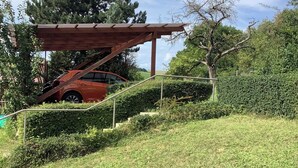 Une voiture rouge est garée sous une pergola en bois, sur une pente herbeuse bordée d’une rambarde en métal.