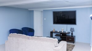 Ein Wohnzimmer mit einem beigefarbenen Sofa, zwei blauen Sesseln, einem flachbildfernseher an der Wand, einem kleinen Tisch mit Dekoration und einer Lampe.