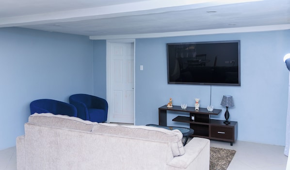 Una sala de estar con un sofá color beige, dos sillones azules, un televisor de pantalla plana montado en la pared, una mesita con objetos decorativos y una lámpara.