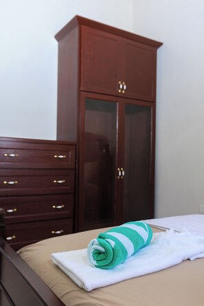 A neatly made bed with a rolled towel, a wooden dresser with drawers, and a wardrobe with glass doors. -    4-bedroom house in marvelous Choiseul with fitness room, AC (Delcer)