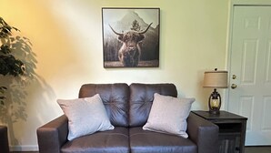 Un sofá de cuero marrón con dos cojines, un cuadro enmarcado de un toro en la pared, una lámpara en una mesita auxiliar y una puerta al fondo.