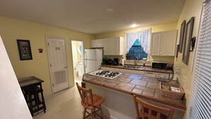 Una cocina compacta con una isla central, dos taburetes de madera, un refrigerador, un fregadero y una ventana con cortinas de encaje.