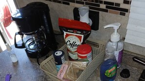 Een aanrecht met een koffiezetapparaat, koffiezakjes, een mand, schoonmaakmiddelen en een spoelbak.
