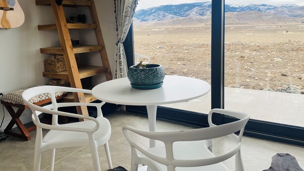 A room with a large window, a wooden ladder, a guitar, a table with a patterned bowl, and two white chairs.