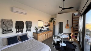 A bedroom with a bed, a desk, and chairs, a kitchenette, and a large window with a view.