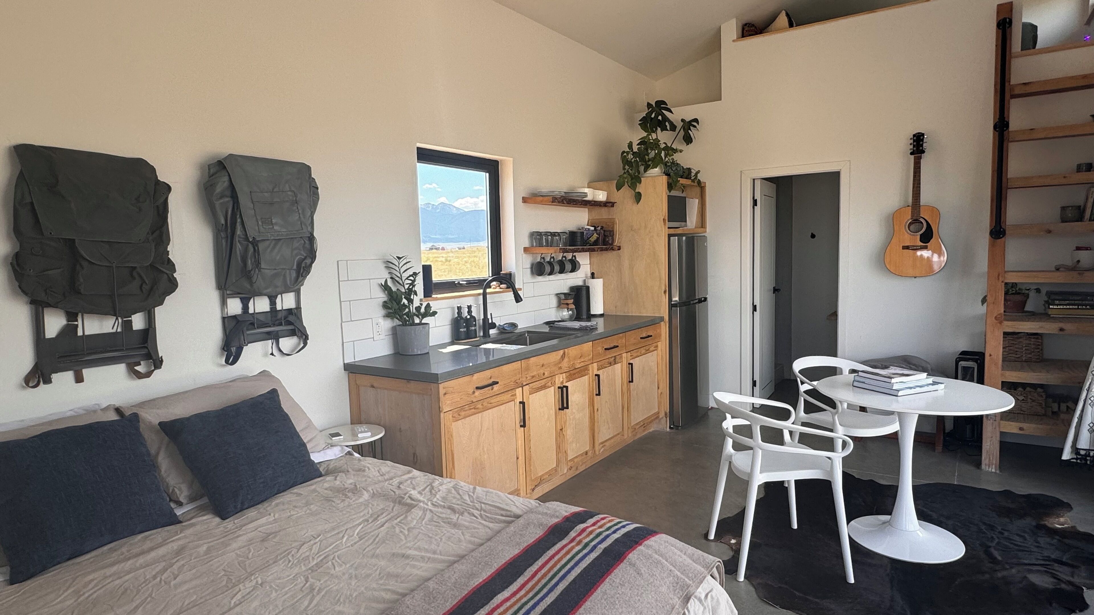 A compact bedroom with a bed, bedside tables, and a view of the outdoors. Adjacent is a small kitchenette with a sink, stove, and cabinets.