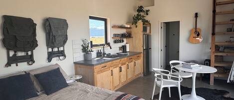 A compact bedroom with a bed, bedside tables, and a view of the outdoors. Adjacent is a small kitchenette with a sink, stove, and cabinets.