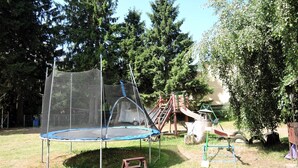 Un trampolino con rete di sicurezza, uno scivolo e un'altalena in un'area erbosa con alberi sullo sfondo.