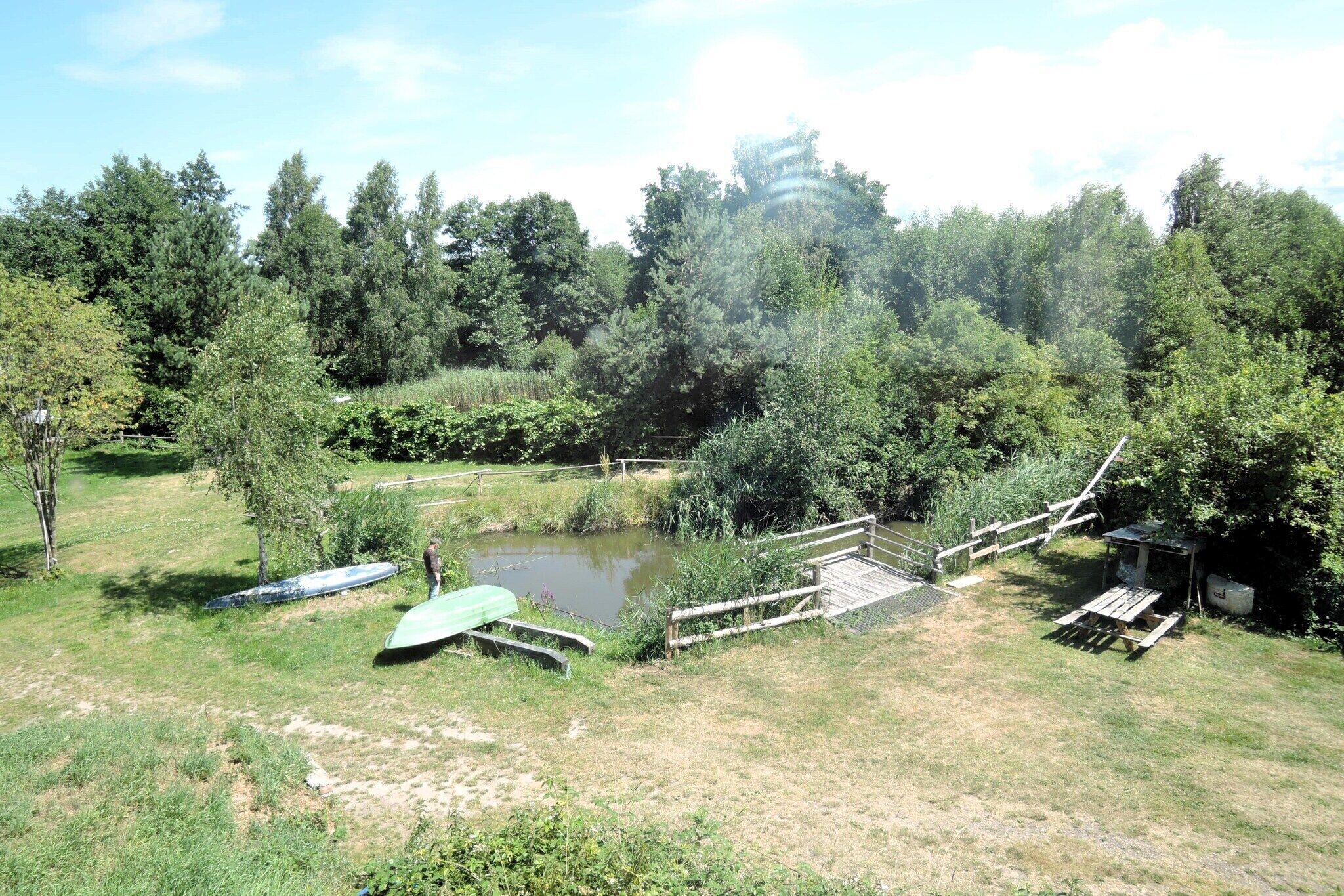 A natural setting with a pond, wooden bridge, picnic table, and a small shelter.