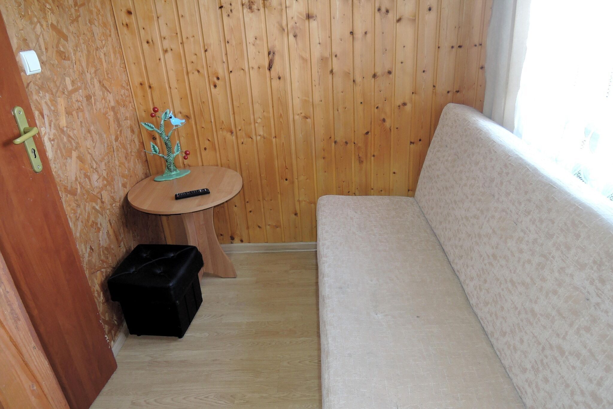 A small room with a wooden wall, a round wooden table with a plant, a black storage box, and a beige sofa.
