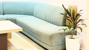 A light blue sectional sofa with a tufted backrest, a white side table, and a potted plant with broad leaves. - Hotel Shri Radha Sneh Mathura (Mathura)