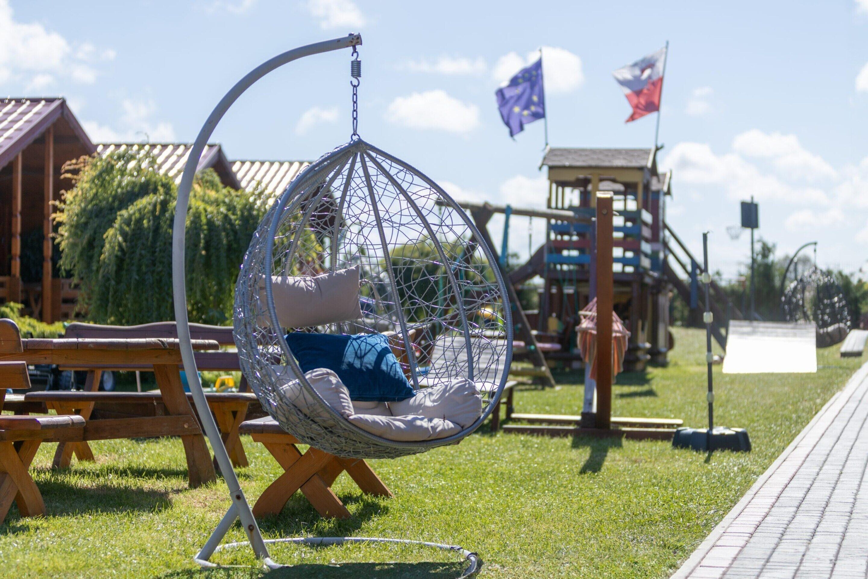 A hanging chair in a garden area with picnic tables and a playground structure in the background.