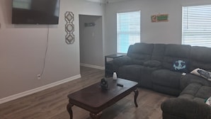 A living room with a sectional sofa, a coffee table, a ceiling fan, a flat-screen TV, and a door labeled "BEACH".