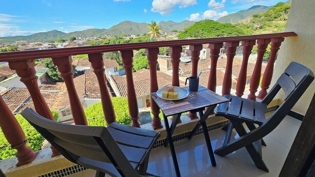A balcony with a table and chairs, overlooking a town with red-roofed buildings and green hills. - Your Retreat in Roldanillo! (Roldanillo)