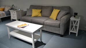 A living room with a grey sofa, two yellow cushions, a white coffee table, a white side table with a book, and a white bedside table with a lamp. - Charming studio in peaceful Long Beach with AC (Long Beach)