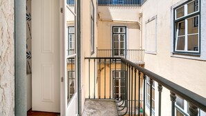 A narrow corridor with a staircase leading to an upper floor. The corridor has a white door and a window on the left. The walls are painted in a light color, and there is a balcony on the upper floor.