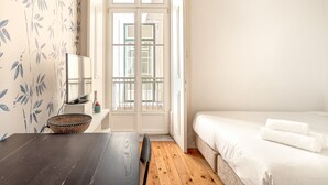 A bedroom with a wooden floor, a bed with white bedding, a dark wooden table, a wicker basket, and a white wall with a floral pattern.