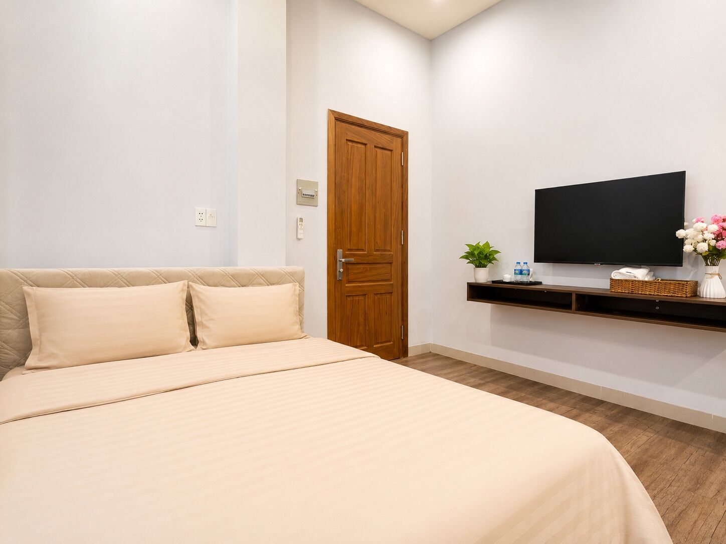 Un dormitorio con una cama, una puerta de madera, un televisor en un estante y una planta pequeña.