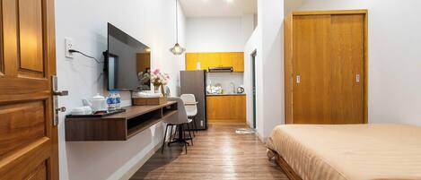 A compact hotel room with a wooden door, a bed, a desk with a chair, a small kitchenette, and a mirror.