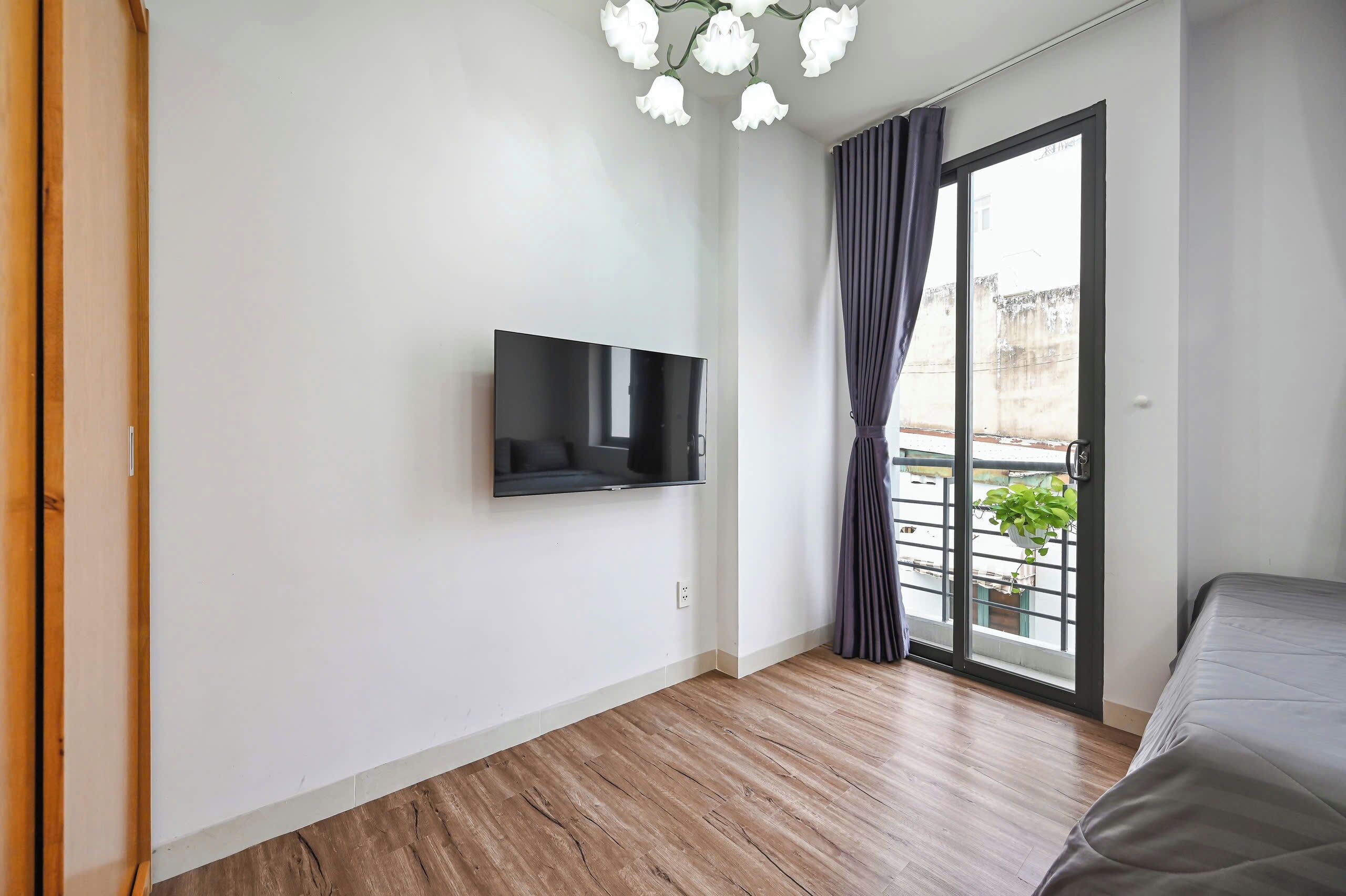 A room with a flat-screen TV mounted on the wall, a sliding glass door to a balcony with a potted plant, and a bed with a grey cover.