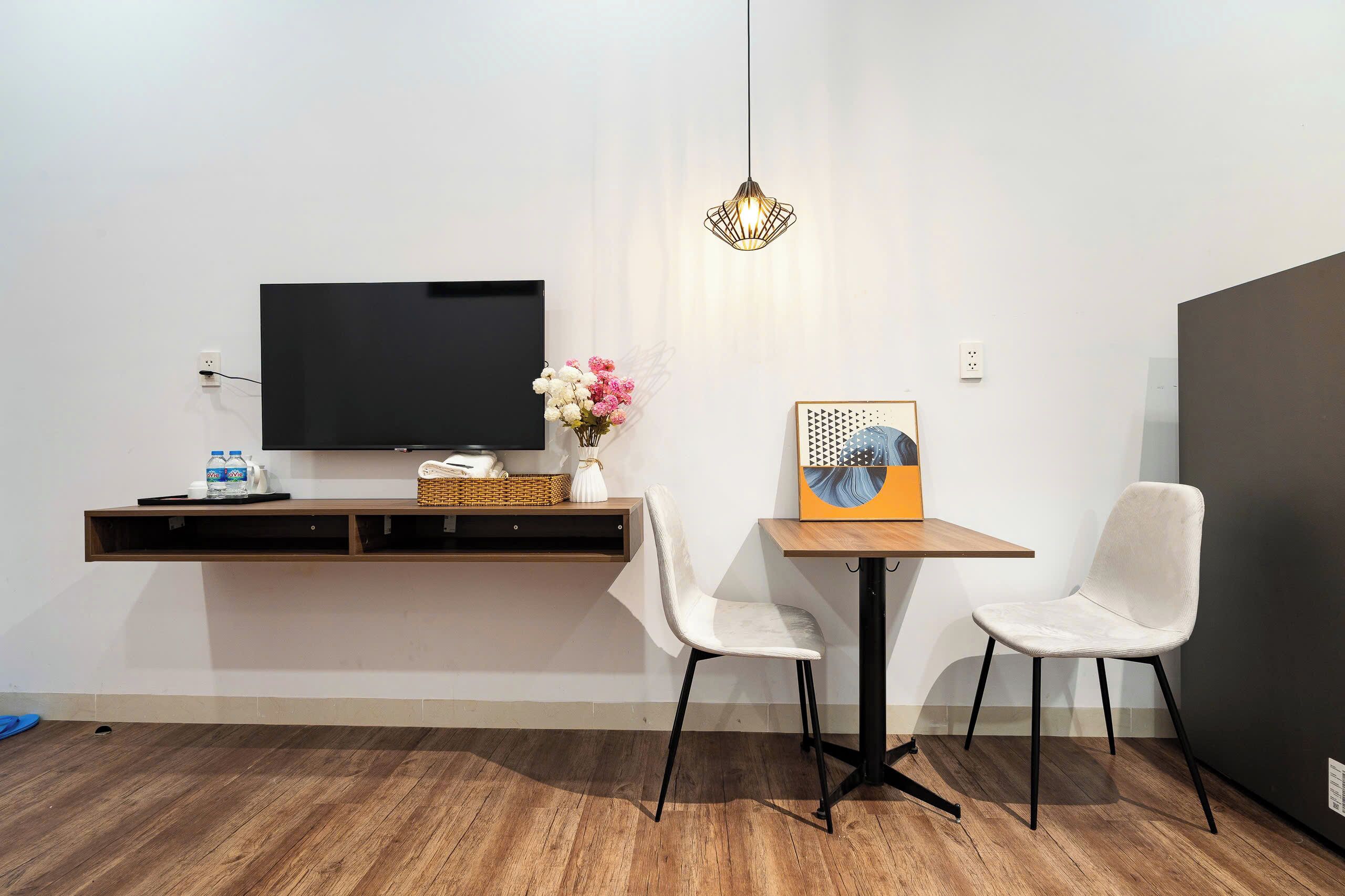 A modern room with a flat-screen TV, a small table with two chairs, a hanging light fixture, and a wall-mounted shelf with a basket and flowers.