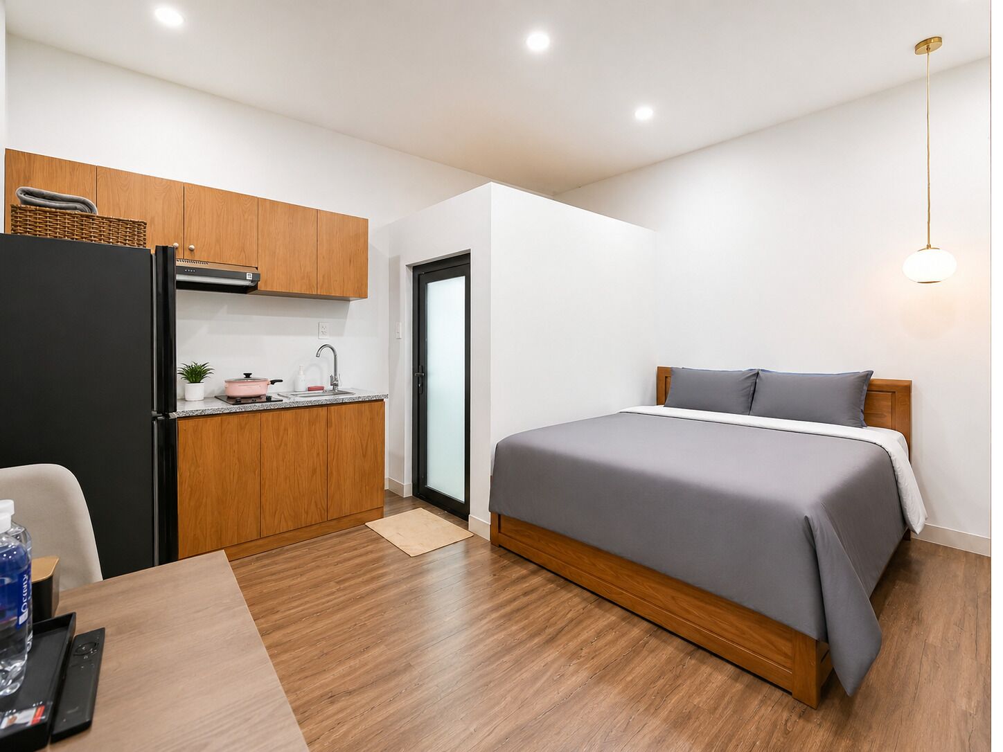 A compact hotel room with a kitchenette, a bed, a wooden floor, and a ceiling light.