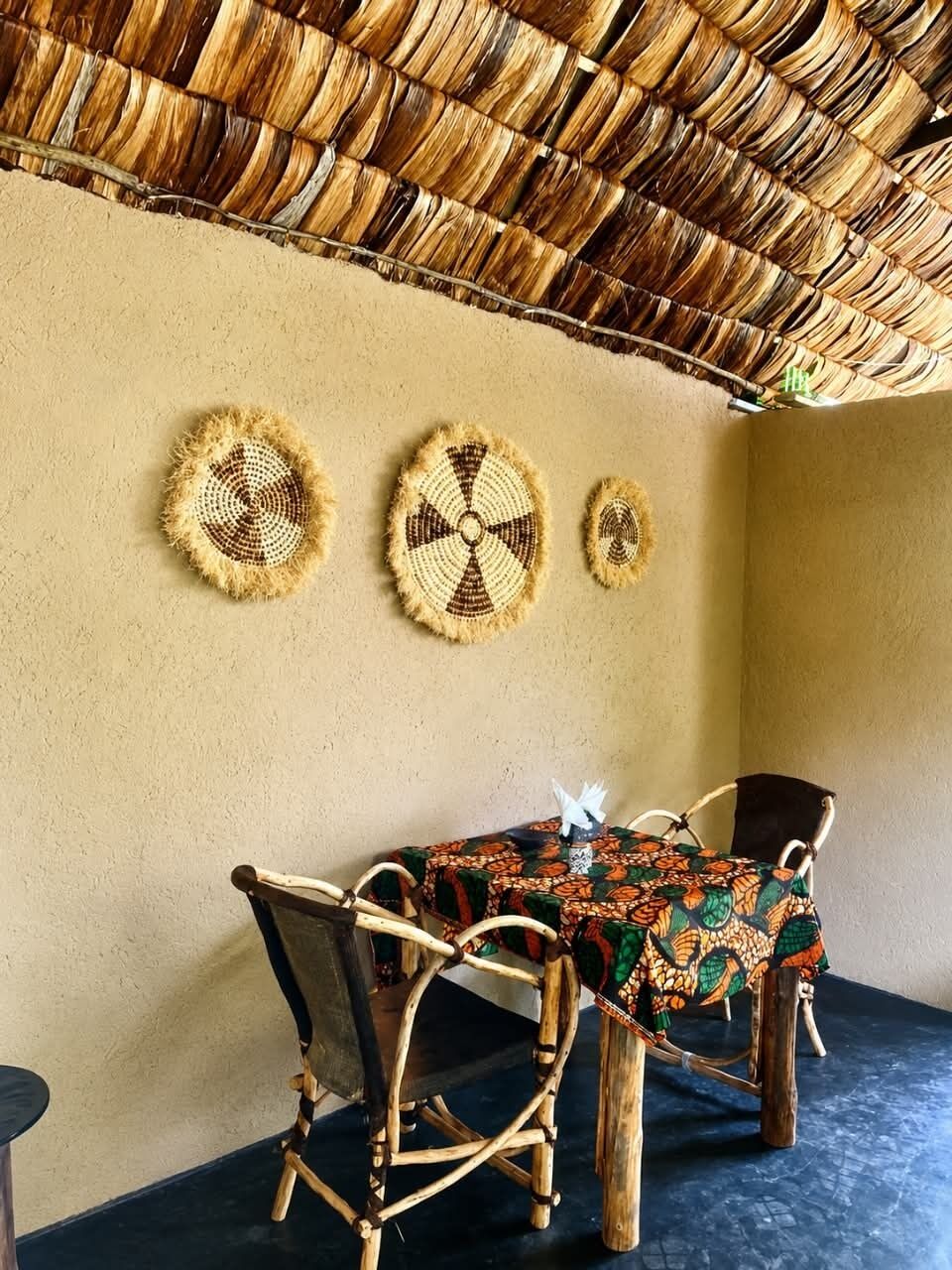Una habitación con techo de paja trenzada, una pared con tres tapices circulares tejidos, y una mesa con mantel estampado.