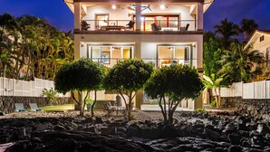 Una casa de dos pisos con balcón, rodeada de vegetación tropical y un litoral rocoso.