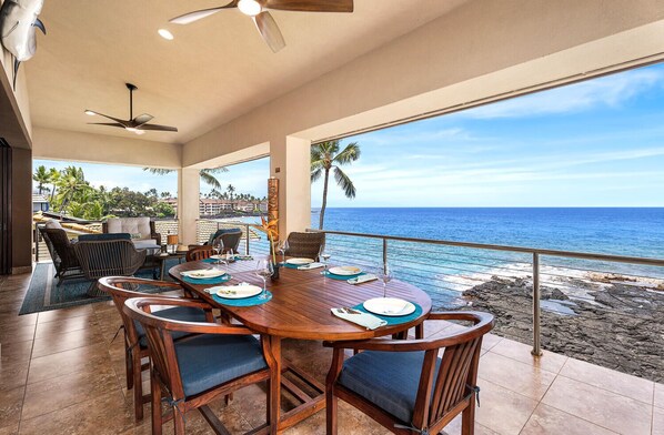 A dining area with a view of the ocean, featuring a wooden table and chairs, a ceiling fan, and a view of the beach.
