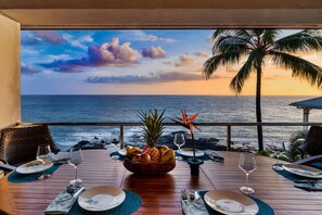 A dining table set with plates, glasses, and a fruit bowl, overlooking a beach with a sunset and palm trees.