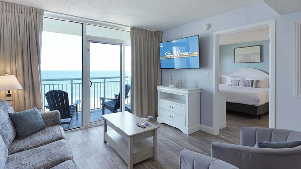A hotel room with a balcony, a bed, a TV, a desk, and a sofa. - Parrot-ise by StayMareni - Oceanfront Suite (Myrtle Beach)