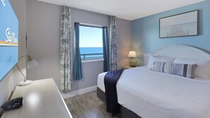 A hotel room with a bed, a desk, a lamp, a window with curtains, and a painting on the wall. - Parrot-ise by StayMareni - Oceanfront Suite (Myrtle Beach)