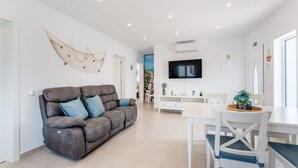 A modern living room with a grey sofa, white dining table, and chairs. There is a TV mounted on the wall, and a hammock decoration on the wall.