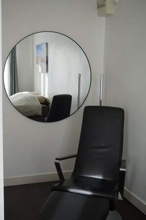 Une pièce comprenant un lit, un fauteuil en cuir noir et un miroir rond accroché au mur.