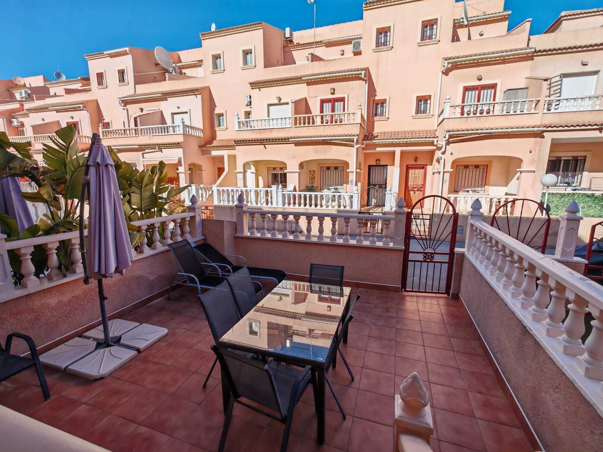 Une terrasse avec une table et des chaises en verre, entourée d’une balustrade blanche, avec des bâtiments en arrière-plan.