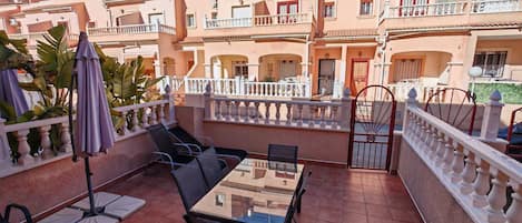 Une terrasse avec une table et des chaises en verre, entourée d’une balustrade blanche, avec des bâtiments en arrière-plan.