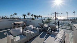 A rooftop terrace with wicker furniture, a table, and a view of the sea and palm trees.