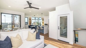 A modern living room with a white sofa, a ceiling fan, and large windows offering a view of palm trees.