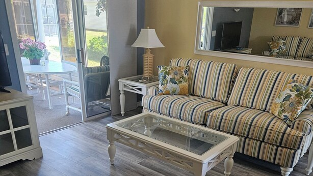 A living room with a striped sofa, a glass coffee table, and a sliding glass door leading to an outdoor patio. - Charming 2-bedroom house in fabulous The Villages with AC (The Villages)