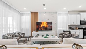 A modern living room with a TV displaying a fireplace, comfortable seating, and a kitchen area.