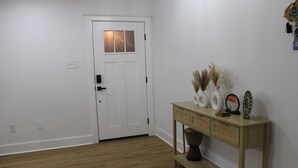 A hallway with a white door, a wooden console table with decorative items, and a light switch on the wall. - Cozy Home in Baton Rouge (Baton Rouge)