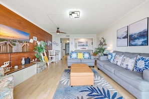 A modern living room with a grey sofa, a wooden accent wall, a ceiling fan, and a large painting of a beach scene.