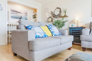A living room with a grey sofa, a blue and yellow patterned pillow, a small table with a lamp, and a decorative mirror on the wall.