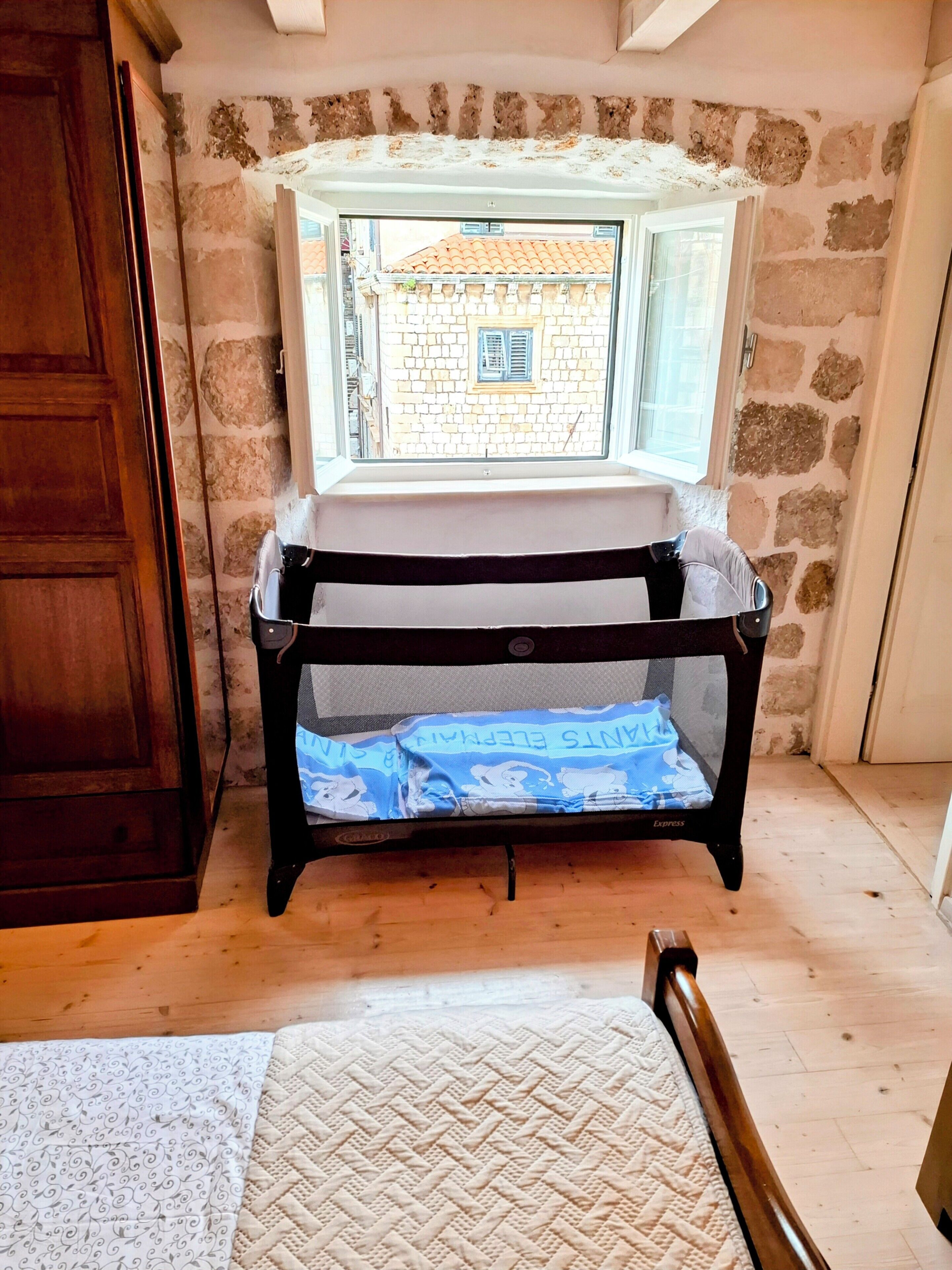 A room with a crib against a stone wall, a window with a view of a building, and a wooden door.