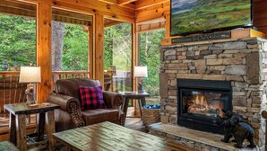 A cabin interior with a stone fireplace, wooden furniture, and large windows offering a view of the forest. - Creekside Dreams – Hot Tub, Fire Pit, Pool Table, 2 Bunk Beds & Creekside Deck (Pigeon Forge)